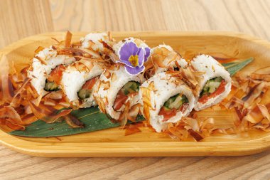 Salatalık ve domatesle dolu suşi ruloları ahşap bir tabakta oturur. Bonito taneleri ve mor çiçeklerle süslenmiş bu yemek taze Japon mutfağını ön plana çıkarıyor..