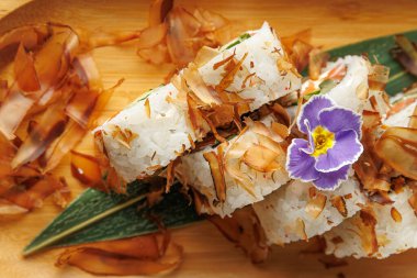 Suşi rulolarının üzerine bonito gevreği ve yenilebilir çiçeklerle süslenmiş çeşitli yiyecekler ahşap bir tahtada servis edilmeye hazırdır..