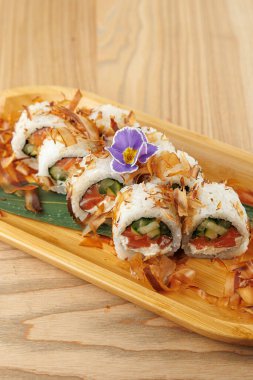 Bir tabakta taze sebze ve balıkla doldurulmuş suşi ruloları, bonito taneleri ve bir çiçek garnitürüyle kaplı. Bu yemek Japon mutfağını ön plana çıkarıyor..