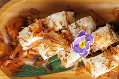 Suşi ruloları ahşap bir tabakta servis edilir. Üstünde bonito taneleri sergilenir. Küçük mor bir çiçek tabağa dekoratif bir dokunuş ekler..