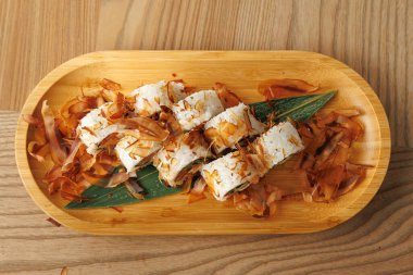 Tahta tabakta suşi ruloları var. Hafif ahşap zemin üzerine yerleştirilmiş bonito gevrekleriyle süslenmiş. Bu yemek Japon mutfağından canlı tatlar sergiliyor..