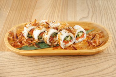 Sebze ve balıklarla dolu suşi ruloları ahşap bir tabakta sunulur. Bonito taneleri tabağın etrafına sade ahşap bir masaya saçılmış..