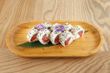 Ahşap bir tabak somonla doldurulmuş ve mor çiçeklerle süslenmiş birkaç suşi rulosu sergiliyor. Düzenleme, bu yemeklerin karmaşık hazırlık ve sunumunu vurguluyor.