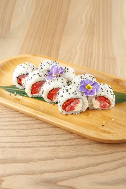 Suşi ruloları taze malzemeler ve renkli dolgular sergiliyor. Her parça ahşap bir tabakta iyi düzenlenmiş ve sunum için canlı çiçeklerle süslenmiş..