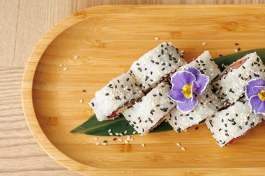 Birkaç suşi rulosu ahşap bir tabakta susam tohumları serpiştirilir ve üzerinde bir çiçek vardır. Tabakta Japon aşçılık ve sunum estetiği karışımı sergileniyor..
