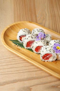 Suşi ruloları taze balıkla doldurulur ve yenilebilir çiçeklerle süslenir ahşap bir tabakta güzelce düzenlenir. Ayarlar yemeğin renkli sunumunu tamamlar.