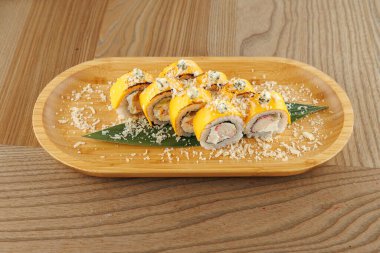 Suşi ruloları yeşil yapraklı ahşap bir tabakta güzelce düzenlenir. Tabakta canlı sarı malzemeler ve parçalanmış malzemeler var..