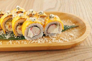 Tabakta düzgün sıralanmış taze suşi ruloları var. Ekmekler dilimlenmiş peynirle kaplanır ve yeşil bir yaprağa konur. Renkli malzemelerini vurgularlar..