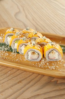 Suşi ruloları ahşap bir tabakta sunulur. Her rulo yengeç eti ve mango, çıtır çıtır malzemelerle süslenmiş ve yeşil garnitürle servis ediliyor..