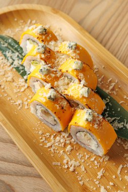 Suşi ruloları ahşap bir tabakta güzelce düzenlenir. Ekmekler lezzetli bir garnitürle kaplanmış ve detaylara özen gösterilmiş..