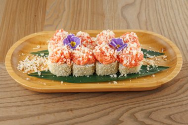 Somon ve ezilmiş malzemelerle kaplı taze suşi ruloları ahşap bir tabakta güzelce düzenlenir. Bir çiçek süslemesi yemeğe biraz renk katar..