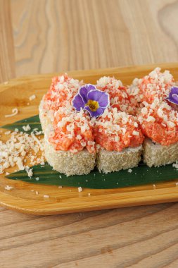 Bir suşi tabağı üzerinde pembe deniz ürünleri ve rendelenmiş malzemelerle küçük pirinç kareleri içerir. Yeşil yapraklı ve mor çiçekli bambu bir tabağa konur..