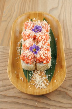 Suşi tabağı, içinde pirinç, taze balık ve malzemeler bulunan ahşap bir tabakta düzenlenir. Tabak bir yaprak ve yenilebilir çiçeklerle sunuluyor, canlı renkler sergileniyor..