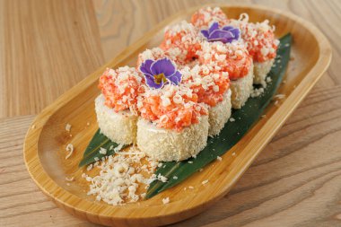 Üzerinde pembe karides ve beyaz taneler olan karidesli suşi ruloları yeşil yapraklı ahşap bir tabağa konur. Yenilebilir çiçekler akşam yemeği için yemeğe renk katar.