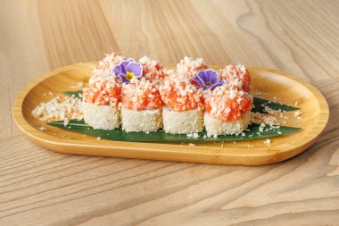 Suşi ruloları ahşap bir tepside pembe deniz ürünleri malzemeleriyle servis edilir ve küçük çiçeklerle süslenir. Bu yemek Japon yemek ortamında lezzetleri ön plana çıkarıyor..