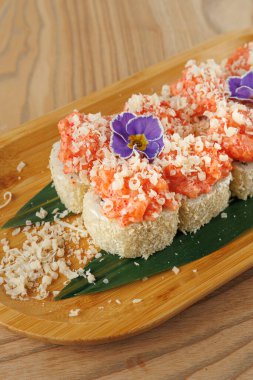 Tahta bir tabakta karides ve öğütülmüş malzemelerle kaplı suşi ruloları sergilenir. Yenilebilir bir çiçek yemeğe renk katar. Ayar, yiyeceklerin canlı sunumunu vurguluyor..