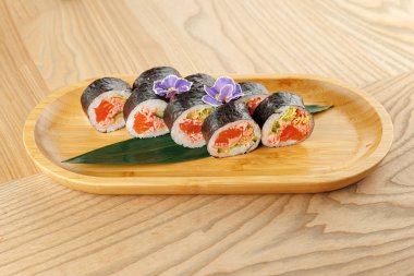 Taze balık ve sebzelerle dolu suşi ruloları bambu tabakta oturur. Tabakta dekoratif çiçekler ve yeşil bir yaprak var. Ortam sade ve sıcak, ahşap bir masa var..