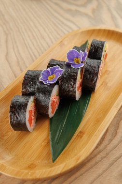 Suşi ruloları ahşap bir tabağa güzelce yerleştirilir. Ekmeğe pirinç ve dolgu malzemesi sarılmış koyu deniz yosunu konuyor. Yenilebilir çiçekler sunuma dekoratif bir dokunuş ekler.