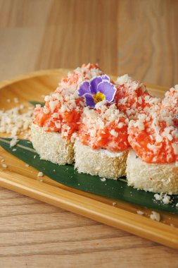Suşi tabağında renkli malzemeler ve ahşap bir tabakta servis edilen dekoratif çiçekler sergileniyor. Yemek, Asya mutfağının zevkine hitap eden doku ve tatların karışımını vurguluyor..