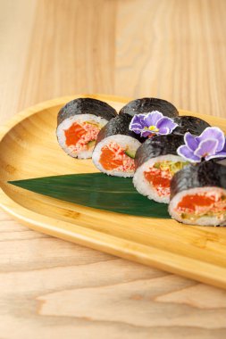 Suşi dürümleri yeşil yapraklı ahşap bir tabakta güzelce düzenlenir. Mor çiçekler renk katar, geleneksel Japon mutfağını sergiler..