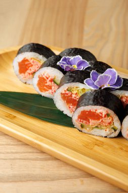 Taze deniz ürünleri ve sebzelerle dolu suşi ruloları ahşap bir tabakta sunulur, mor yenilebilir çiçeklerle süslenir, canlı renkler ve dokular sergilenir..