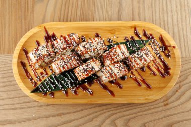 Suşi rulosu koleksiyonu ahşap bir tabakta düzenlenir. Ekmekler susam tohumlarıyla kaplı ve koyu bir sos serpiştirilmiş. Ayar yemeğin sunumunu vurguluyor.