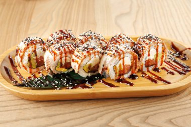 Ahşap bir tabakta renkli suşi ruloları serpiştiriliyor, üzerine sos serpiştirilmiş ve susam tohumları serpiştirilmiş. Yemek, lokantaları öğle yemeğinde lezzetli bir yemek yemeye davet ediyor..