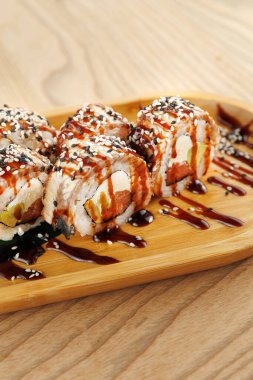 Balık ve sebzelerle dolu suşi ruloları ahşap bir tabakta serpiştirilir, üzerine sos serpiştirilir ve susam tohumları serpilir. Sahne Japon mutfağına odaklanan bir yemek deneyimini gösteriyor..
