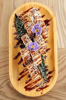 Sushi tohumu ve sosla kaplanmış suşi ruloları yeşil yapraklar ve dekoratif çiçek aksanıyla çevrili ahşap bir tabakta sergileniyor..