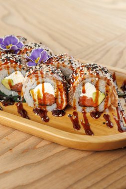 Somon ve krem peynirle dolu suşi ruloları ahşap bir tabakta düzenlenir. Tabak sosla kaplandı ve yenilebilir çiçeklerle süslendi. Renkli bir sunum sunuldu..