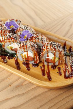 Suşi ruloları ahşap bir tabağa konur. Ekmekler susam tohumlarıyla kaplı ve sosla ıslatılmış. Küçük bir çiçek tabağa biraz süs katar..