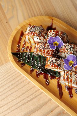 Suşi dürümleri ahşap bir tabakta, sos ve yenilebilir çiçeklerle süslenir. Sahne canlı renkler ve restoran ortamında taze malzemeler yakalıyor..