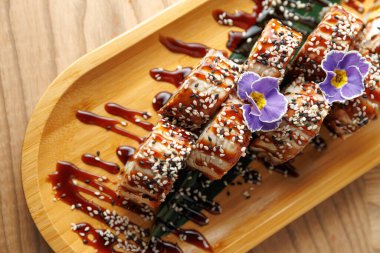 Suşi ruloları ahşap bir tepside üzerine sos serpilmiş ve susam tohumları serpiştirilmiş. Yenilebilir çiçekler dekoratif bir dokunuş ekler. Yemek masasında taze suşinin tadını çıkarıyorum..