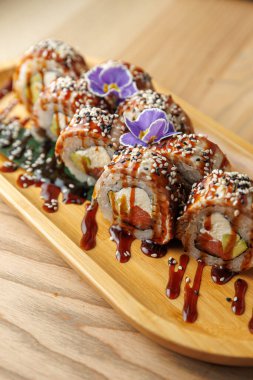 Suşi dürümleri ahşap bir tabakta, sos ve yenilebilir çiçeklerle süslenir. Bu yemek, bir yemek yerinde lezzet ve dokuların karışımını sergiliyor..