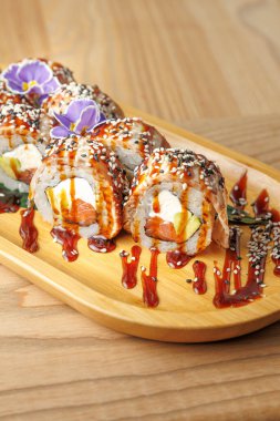 Suşi rulosu seçimi ahşap bir tabakta düzenlenir. Ekmekler malzemelerle dolu, sosla kaplı ve dekorasyon için küçük çiçekler..