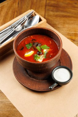 Zengin bir kase çorba ahşap bir masanın üzerine konur. Çorba sebze ve bitki parçalarını içeriyor. Küçük bir tabak krema kâsenin yanında servis edilir..