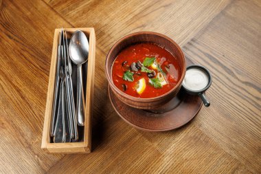 Bir kase kırmızı çorba, içinde çatal bıçak bulunan bir konteynırın yanında ahşap bir masanın üzerinde oturur. Çorbada bitkiler ve limon dilimleri var. Yakınlarda bir fincan krema var..