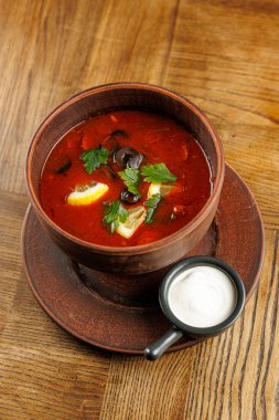 Pancar ve sebzelerle dolu, otlar ve limon dilimleriyle süslenmiş canlı kırmızı bir çorba kasesi. Küçük bir tabak ekşi kremayla birlikte ahşap bir masada servis edilir..