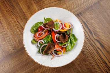 Bir kasede taze yeşillik, olgun domates ve dilimlenmiş soğandan yapılmış bir salata bulunur. Et parçaları doku ekliyor. Tabak ahşap bir masaya konur, doğal malzemelerin altını çizer..