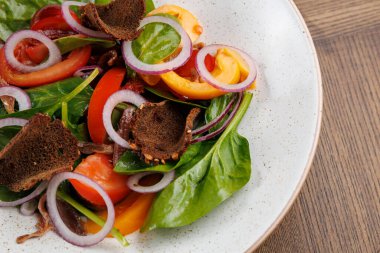 Bir kase taze salata ıspanak, dilimlenmiş domates ve düzgün sıralanmış kırmızı soğan içerir. Salata siyah ekmek parçalarıyla kaplı ve ahşap bir masada servis ediliyor..