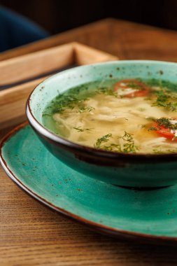 Sebzeli ve otlu tavuk çorbası servisi ahşap bir masada mavi bir kaseye konur. Restoran manzarası rahat ve davetkar. Öğle yemeği sırasında sıcak bir yemeğin tadını çıkarmak için mükemmel..