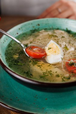 Tahta bir masada bir kase çorba servis edilir. Çorbada sebze parçaları, domates dilimleri ve haşlanmış yumurta var. Taze bitkiler tabağa serpilmiş..