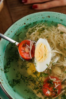 Bir kase tavuk çorbası erişte, domates ve haşlanmış yumurta içerir. Taze dereotu, lezzet katarak yemeği süslüyor. Ayar, kırsal bir ahşap masayı vurguluyor..