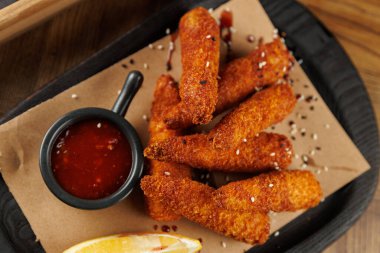 Bir tabakta düzgün sıralanmış kızarmış atıştırmalıklar var. Kırmızı sosla dolu küçük siyah bir kase aperatiflerin yanında oturur. Limon dilimi taze bir dokunuş katar..