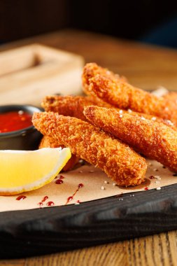 Kızarmış çubuklar ahşap bir tahtanın üzerinde, limon dilimi ve sos ile servis edilir. Sahne, sıcacık bir yemek atmosferi yakalıyor..