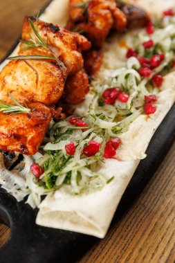 Izgara tavuk şişleri yanında doğranmış otlar, soğanlar ve nar çekirdekleri ile birlikte yassı bir ekmeğin üzerinde servis edilir. Bu yemek canlı renkler ve taze malzemeler sergiliyor..