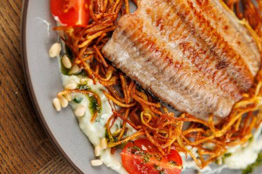 Izgara balık, taze domatesler ve yuvarlak bir tabakta otlarla süslenmiş kızarmış eriştelerden oluşan bir yatağa yerleştirilir. Ayar, farklı mutfaklardan çeşitli besin unsurlarını vurguluyor..