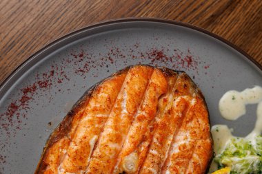 Izgara somon bir porsiyon baharatla süslenmiş siyah bir tabakta oturur. Taze sebzeler yemeğe eşlik eder, aroma ve dokuların bir karışımını sergiler..