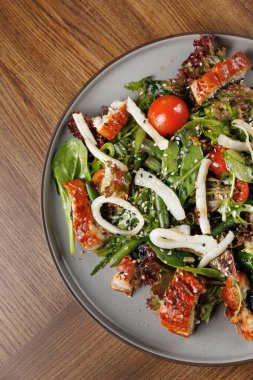 Bir kasede yeşil yapraklı taze bir salata, kiraz domatesleri ve ahşap bir masaya dizilmiş ızgara balık parçaları var. Lezzetli bir yemek deneyimini davet ediyor..