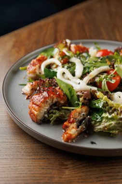 Izgara yılan balığı salatası yuvarlak bir tabakta duruyor. Tabakta yeşiller, dilimlenmiş domatesler ve taze sebzeler yer alıyor. Ortam sıcak ve davetkar, yemek yemek için mükemmel..